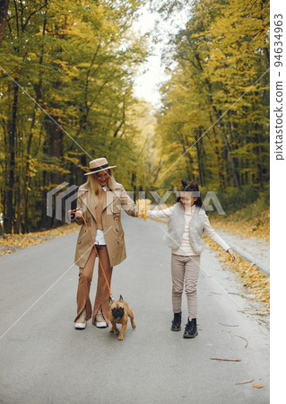 Woman, little girl and dog walking in the autumn park. Brown french bulldog with female owners spend a day at the park playing and having fun. Woman wearing beige coat and a hat. 94634963