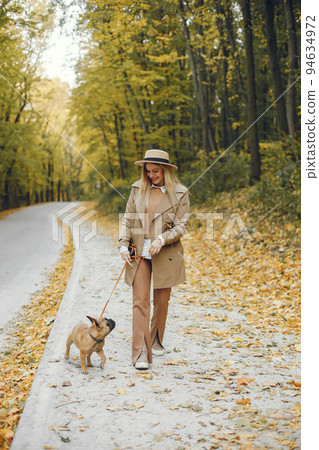 Woman and dog play and have fun in the autumn park. Brown french bulldog with female owner spend a day at the park walking. Woman wearing beige coat and a hat. Woman and dog play and have fun in the autumn park. Brown french bulldog with female owner spend a day at the park walking. Woman wearing beige coat and a hat. 94634972