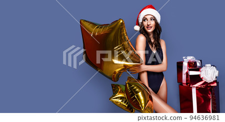 Portrait of young, sexy and beautiful woman in swimsuit and Santa hat, holding golden baloon, near gift boxes. Christmas, xmas, x-mas and winter concept 94636981