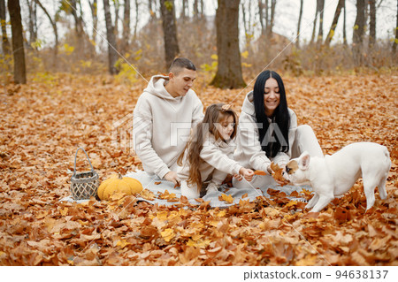 Man, woman, their little girl and dog sitting on a blanket in autumn forest. Brunette woman and man play with daughter. Family wearing beige sportive costumes. Man, woman, their little girl and dog sitting on a blanket in autumn forest. Brunette woman and man play with daughter. Family wearing beige sportive costumes. 94638137