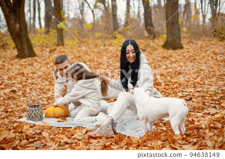 Man, woman, their little girl and dog sitting on a blanket in autumn forest. Brunette woman and man play with daughter. Family wearing beige sportive costumes. 94638149