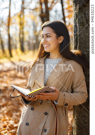Beautiful woman reading a book in autumn park. Young girl standing near tree with a book. Brunette woman wearing beige coat. 94638223