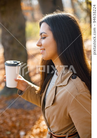 Beautiful woman drink coffee and posing for the camera in autumn park. Young girl standing near tree with a coffee. Brunette woman wearing beige coat. Beautiful woman drink coffee and posing for the camera in autumn park. Young girl standing near tree with a coffee. Brunette woman wearing beige coat. 94638224