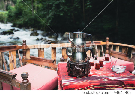 Turkish tea chrome kettle and glasses on a restaurant table with chairs near a river in an outdoor Turkish tea chrome kettle and glasses on a restaurant table with chairs near a river in an outdoor 94641403