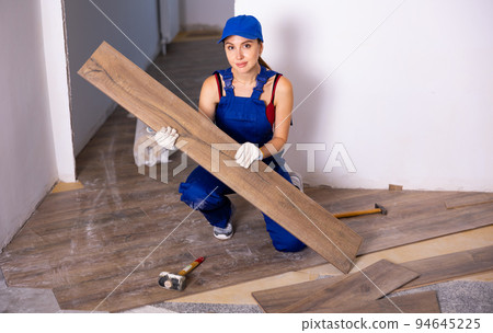 Portrait of beautiful young woman in work clothes and cap installing new wooden laminate flooring sitting squat 94645225