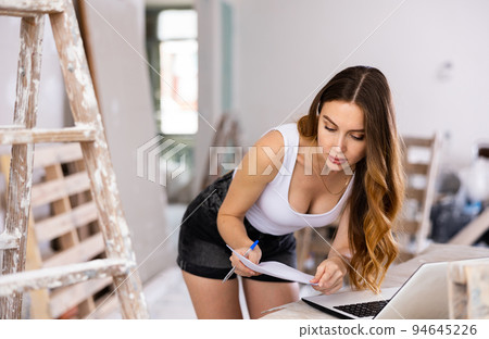 Young attractive girl checks the quality and correctness of the completed repairs using a laptop inside cottage Young attractive girl checks the quality and correctness of the completed repairs using a laptop inside cottage 94645226