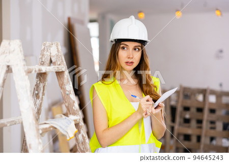 Female supervisor in safety vest and hard hat standing with papers at construction site Female supervisor in safety vest and hard hat standing with papers at construction site 94645243