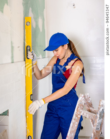 Woman builder in blue overalls measures deviations from a given vertical wall using building level 94645347