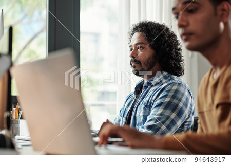 Pensive Indian programmer with curly hair looking at computer screen 94648917