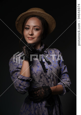 The middle plan is a portrait of a medieval young woman in a blue vintage dress with a black lace collar and a hat,posing on a dark background.Historical fashion of the 19th century.High quality photo 94648974