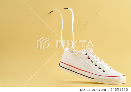 White female sport sneakers with flying shoe lace against yellow background, Casual trendy shoes closeup, Creative minimal design with copy space 94651330