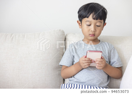An adorable happy Asian boy wearing a gray shirt and blue-white striped shorts is having fun playing with his smartphone on a cream sofa. looking at the mobile screen. An adorable happy Asian boy wearing a gray shirt and blue-white striped shorts is having fun playing with his smartphone on a cream sofa. looking at the mobile screen. 94653073