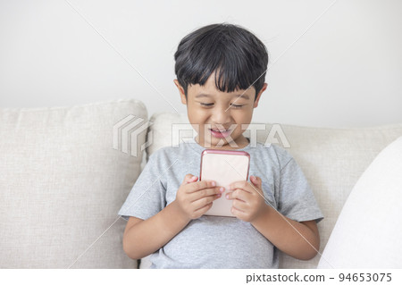 An adorable happy Asian boy wearing a gray shirt and blue-white striped shorts is having fun playing with his smartphone on a cream sofa. looking at the mobile screen. An adorable happy Asian boy wearing a gray shirt and blue-white striped shorts is having fun playing with his smartphone on a cream sofa. looking at the mobile screen. 94653075