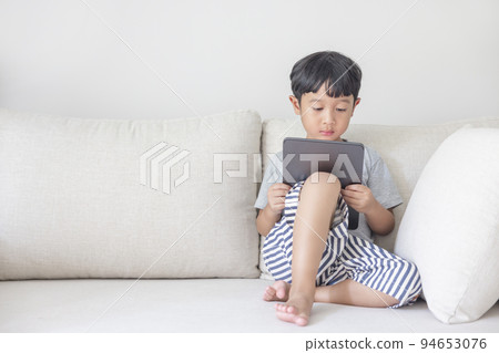 Adorable happy Asian boy wearing a gray shirt and blue-white striped shorts is having fun playing with his tablet on a cream sofa. looking at the mobile screen. Adorable happy Asian boy wearing a gray shirt and blue-white striped shorts is having fun playing with his tablet on a cream sofa. looking at the mobile screen. 94653076