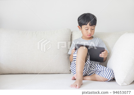 Adorable happy Asian boy wearing a gray shirt and blue-white striped shorts is having fun playing with his tablet on a cream sofa. looking at the mobile screen. Adorable happy Asian boy wearing a gray shirt and blue-white striped shorts is having fun playing with his tablet on a cream sofa. looking at the mobile screen. 94653077