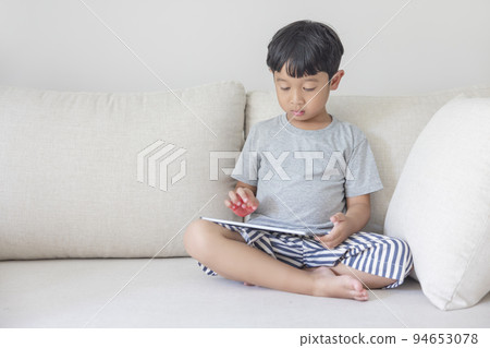 Adorable happy Asian boy wearing a gray shirt and blue-white striped shorts is having fun playing with his tablet on a cream sofa. looking at the mobile screen. Adorable happy Asian boy wearing a gray shirt and blue-white striped shorts is having fun playing with his tablet on a cream sofa. looking at the mobile screen. 94653078