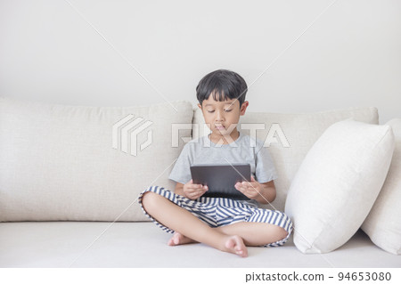 Adorable happy Asian boy wearing a gray shirt and blue-white striped shorts is having fun playing with his tablet on a cream sofa. looking at the mobile screen. Adorable happy Asian boy wearing a gray shirt and blue-white striped shorts is having fun playing with his tablet on a cream sofa. looking at the mobile screen. 94653080