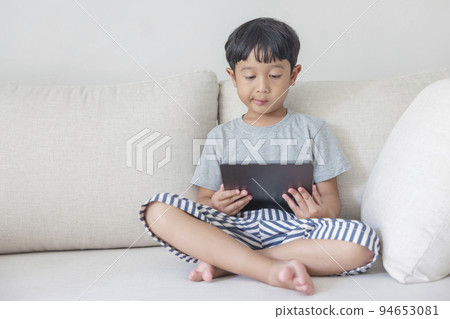 Adorable happy Asian boy wearing a gray shirt and blue-white striped shorts is having fun playing with his tablet on a cream sofa. looking at the mobile screen. Adorable happy Asian boy wearing a gray shirt and blue-white striped shorts is having fun playing with his tablet on a cream sofa. looking at the mobile screen. 94653081