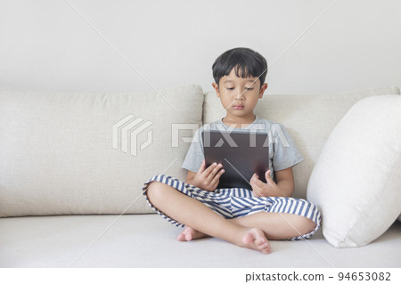 Adorable happy Asian boy wearing a gray shirt and blue-white striped shorts is having fun playing with his tablet on a cream sofa. looking at the mobile screen. Adorable happy Asian boy wearing a gray shirt and blue-white striped shorts is having fun playing with his tablet on a cream sofa. looking at the mobile screen. 94653082