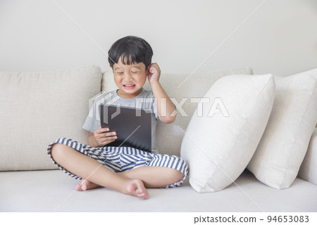 Adorable happy Asian boy wearing a gray shirt and blue-white striped shorts is having fun playing with his tablet on a cream sofa. looking at the mobile screen. Adorable happy Asian boy wearing a gray shirt and blue-white striped shorts is having fun playing with his tablet on a cream sofa. looking at the mobile screen. 94653083