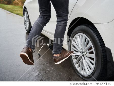 Close up of male auto mechanic standing on metal wrench while unscrewing lug nuts on car wheel. Young man using lug nut wrench while changing flat tire on the road. Concept of emergency road service. 94653140