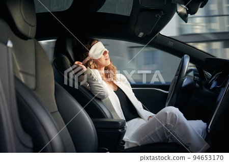 Woman in sleeping mask taking nap in electric car with vehicle autopilot system. Stylish businesswoman sleeping in electro car while using driver-assistance system. Woman in sleeping mask taking nap in electric car with vehicle autopilot system. Stylish businesswoman sleeping in electro car while using driver-assistance system. 94653170