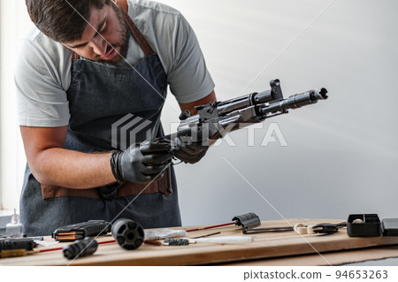 Close up of young man in apron disassembling a gun above the table 94653263