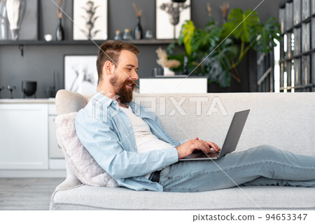 Young bearded man sitting on sofa at home using laptop computer Young bearded man sitting on sofa at home using laptop computer 94653347