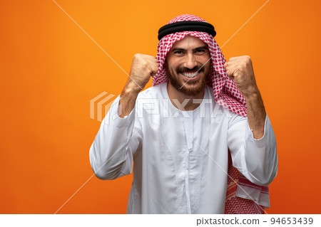 Successful smiling young Arab man celebrating victory on orange background 94653439