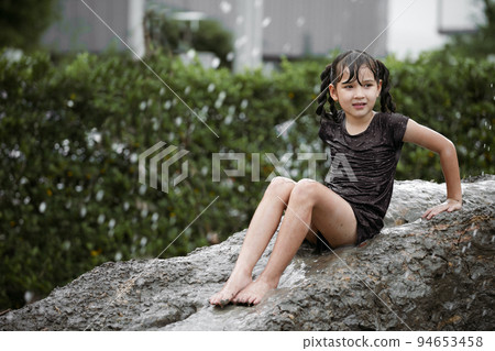 Cheerful child girl playing on heap of wet soil during raining in rainy season. 94653458