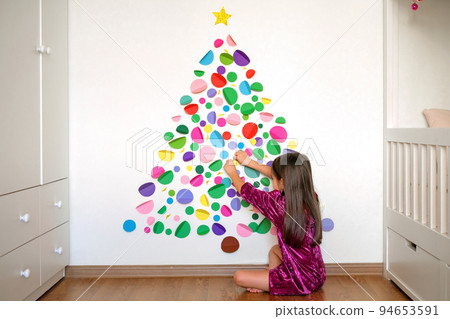 The girl decorates the wall in the nursery with paper multicolored circles that make up the shape of a Christmas tree. An alternative Christmas tree made by handmade The girl decorates the wall in the nursery with paper multicolored circles that make up the shape of a Christmas tree. An alternative Christmas tree made by handmade 94653591