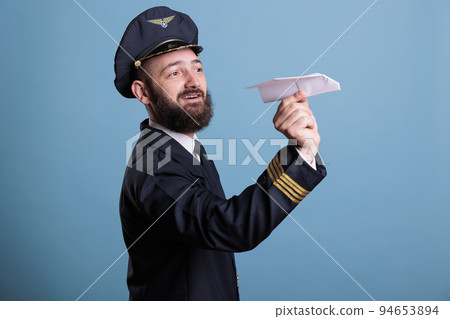 Pilot in professional uniform launching paper plane, holding jet model in hands front view. Aviation academy aviator, aircraft captain in jacket and hat on blue background 94653894