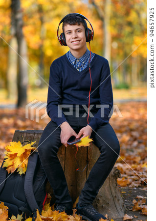 a teenage boy sits in autumn park and listens to music with headphones, trees with yellow leaves on a bright sunny day 94657925