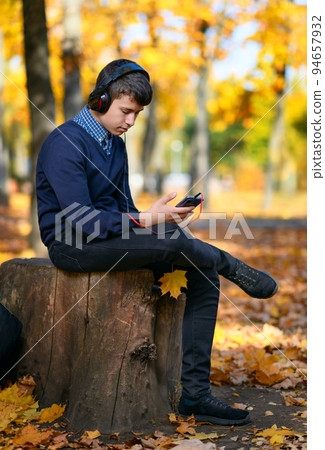 a teenage boy sits in autumn park and listens to music with headphones, trees with yellow leaves on a bright sunny day a teenage boy sits in autumn park and listens to music with headphones, trees with yellow leaves on a bright sunny day 94657932