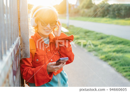 Smiling stylish woman in bright clothes and sunglasses wearing wireless headphones on her head and seting phone for listening music. Walk in city park on sunset. Hipster lifestyle. Selective focus 94660605