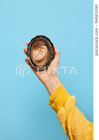 Close-up of female ear with gemstone earrings. Female hand holding small round mirror with reflection of girl's ear isolated over blue background. 94661090