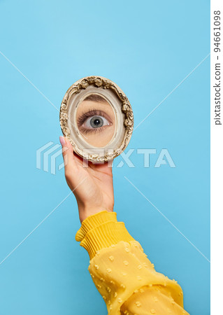 Eyesight. Female hand holding small round mirror with reflection of girl's eye isolated over blue background. Concept of style, beauty, art 94661098