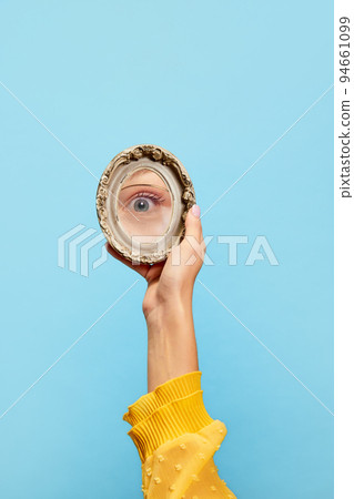 Eyesight. Female hand holding small round mirror with reflection of girl's eye isolated over blue background. Concept of style, beauty, art 94661099
