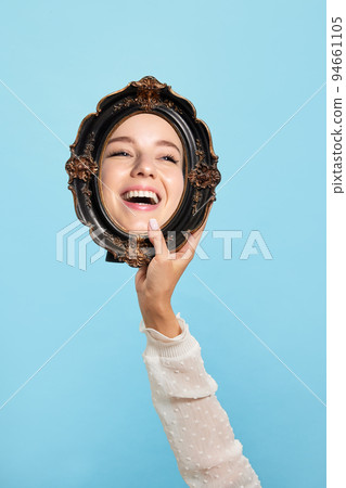 Cheerful girl. Female hand with retro style mirror with reflection of beautiful girl's face isolated over blue background. Concept of emotions, beauty 94661105