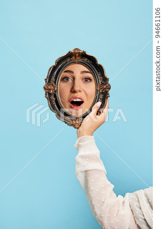 Surprised girl. Woman's hand with retro style mirror with reflection of beautiful girl's face isolated over blue background. Concept of emotions, beauty 94661106