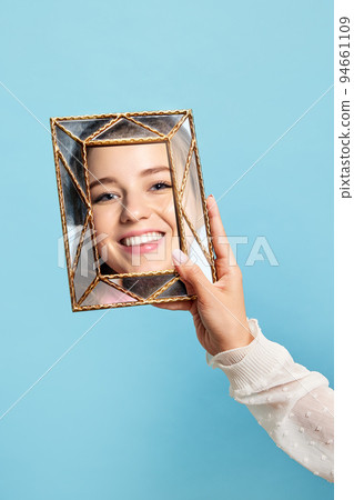 Happy girl. Woman's hand with retro style mirror with reflection of beautiful girl's face isolated over blue background. Concept of emotions, beauty 94661109
