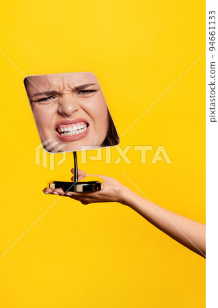 Angry. Female hand holding mirror with reflection of angry girl's face isolated over yellow background. Emotions, facial expressions 94661133