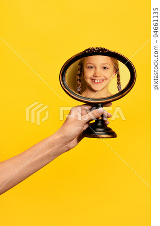 Female hand holding mirror with reflection of smilling little girl isolated over yellow background. Concept of vintage, retro style, beauty, art, creativity 94661135