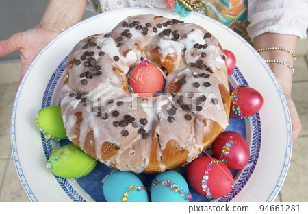 Festive Easter cake prepared for a religious holiday, surrounded by traditionally brightly colored chicken eggs on a round dish held by women's hands. Festive Easter cake prepared for a religious holiday, surrounded by traditionally brightly colored chicken eggs on a round dish held by women's hands. 94661281