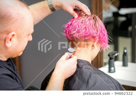 Male hairdresser shaves neck of a young caucasian woman with a short pink hairstyle by electric shaver in a hairdresser salon, close up 94662322