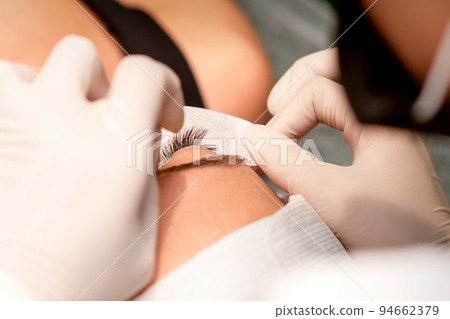 The hands of the cosmetologist are gluing white tape under the eye of the young caucasian woman during the eyelash extension procedure, closeup 94662379