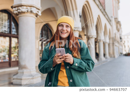 Beautiful stylish woman in hat walking on Market Square in Krakow on autumn day and holding mobile phone. Phone Communication. Urban lifestyle concept. Check social networks, send sms. 94663707