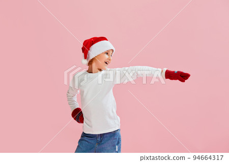 Dance. Cheerful little girl, child in casual style clothes, knitted mittens and Christmas hat having fun over pink background. 94664317
