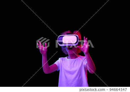 School age girl wearing white t-shirt enjoying 3D technology in virtual reality headset isolated on dark background in neon light 94664347
