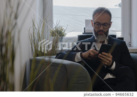 Businessman with beard and glasses uses a smartphone sitting on sofa in office Businessman with beard and glasses uses a smartphone sitting on sofa in office 94665632